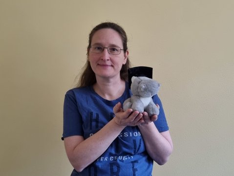 A woman holding a stuffed hippo. She is smiling. The hipp wears a doctorate hat.