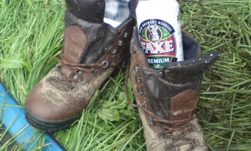 Photo of a pair of muddy boots in the entrance to a tent. There is a can of beer in one bootand a botle of water in the other.