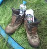 Photo of a pair of muddy boots in the entrance to a tent. There is a can of beer in one bootand a botle of water in the other.