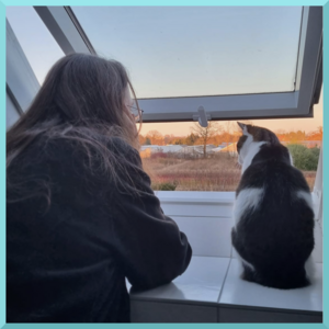 Photo von einer Frau und einer Katze, Rückansicht. Die Frau lehnt auf der Fensterbank eines Dachfensters, die Katze sitzt auf der Fensterbank. Das Dachfenster ist geöffnet und beide schauen hinaus.