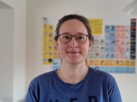 A woman smiling and looking into the camera. In the blurred background there is a poster of the periodic table of the elements on an off white wall.